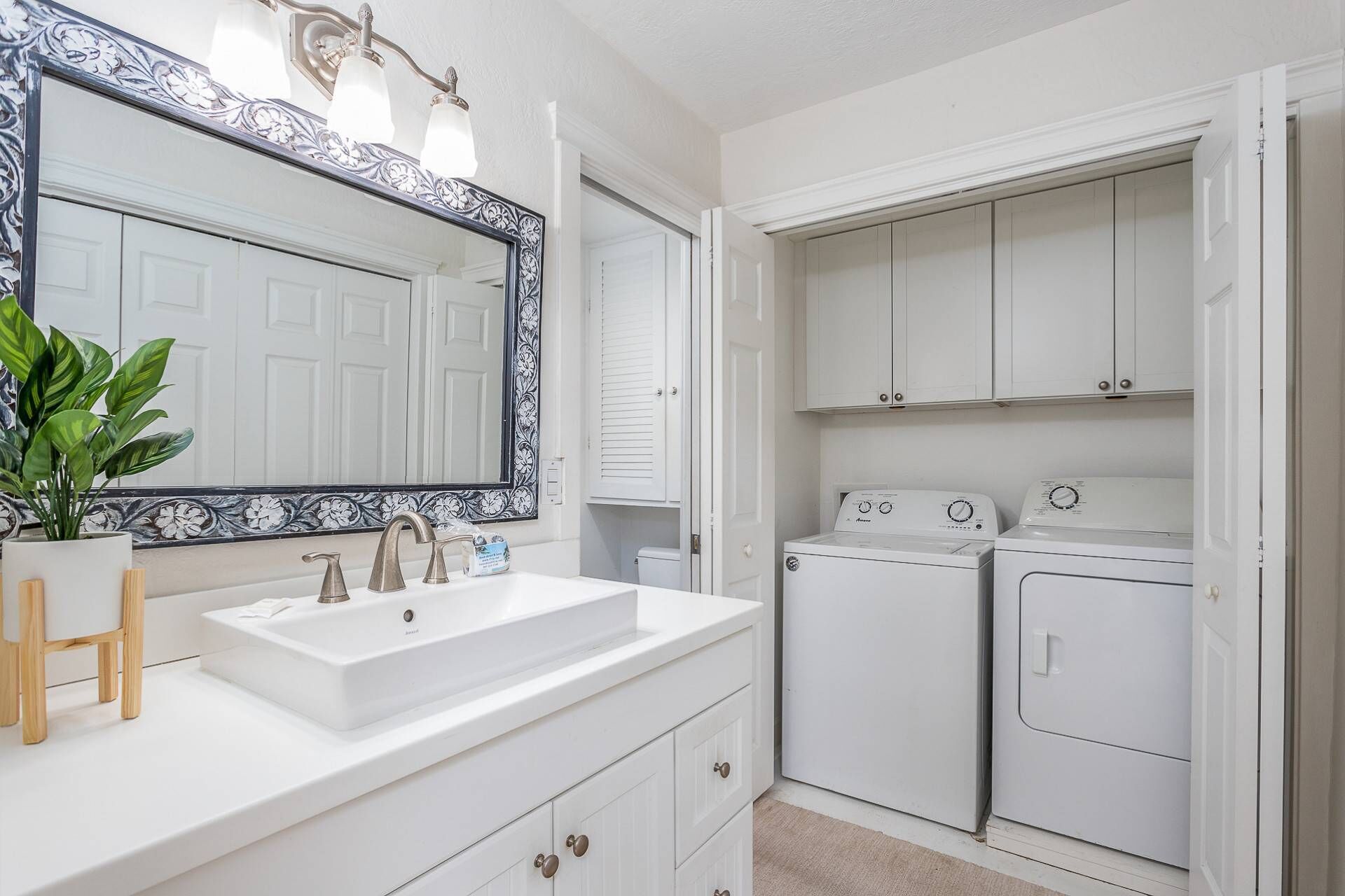 A bathroom with a white sink, two faucets, a mirror with a decorative frame, a potted plant, and a set of white cabinets with a washing machine.
