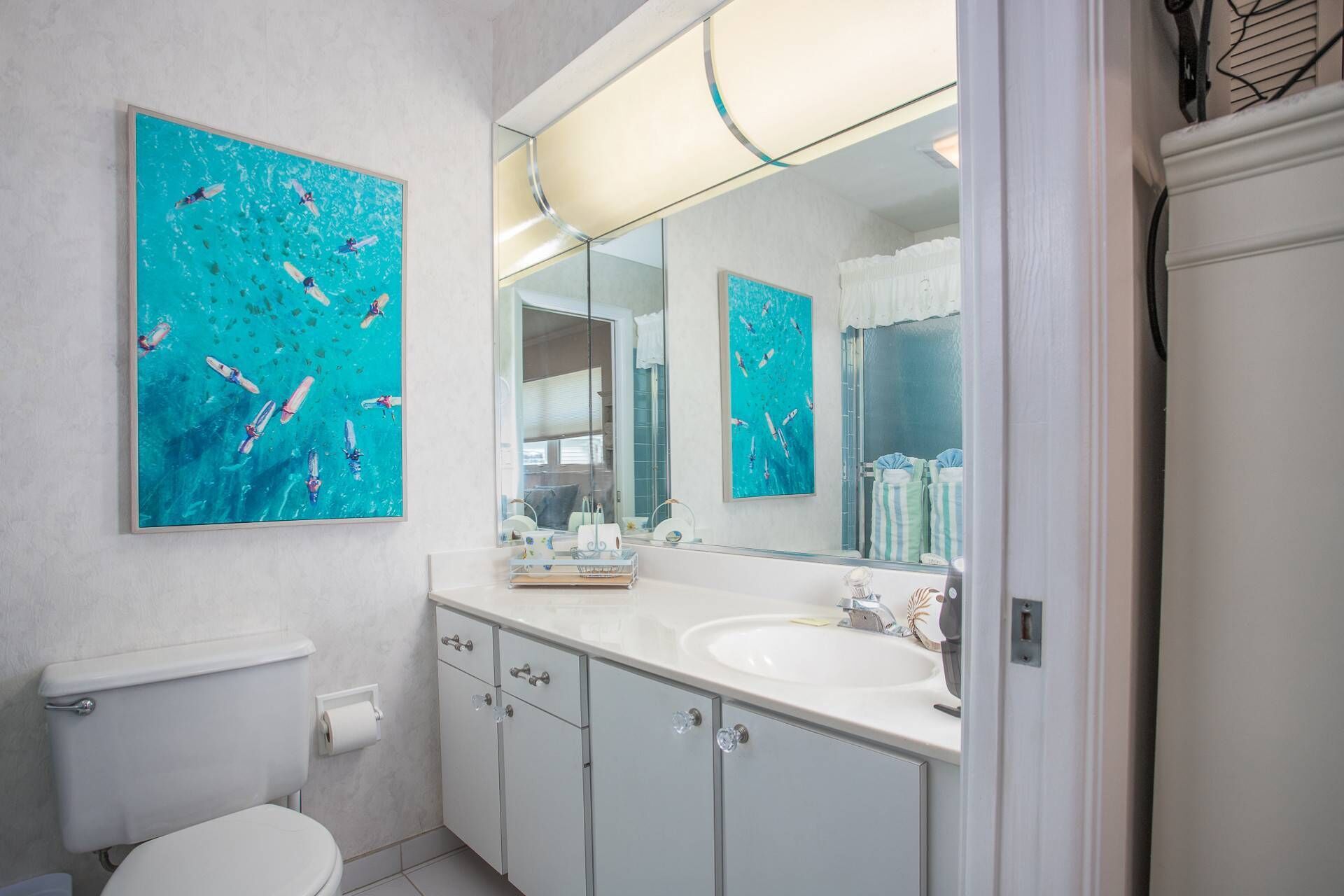A bathroom with a large mirror, two wall-mounted paintings, a white sink, and a toilet.