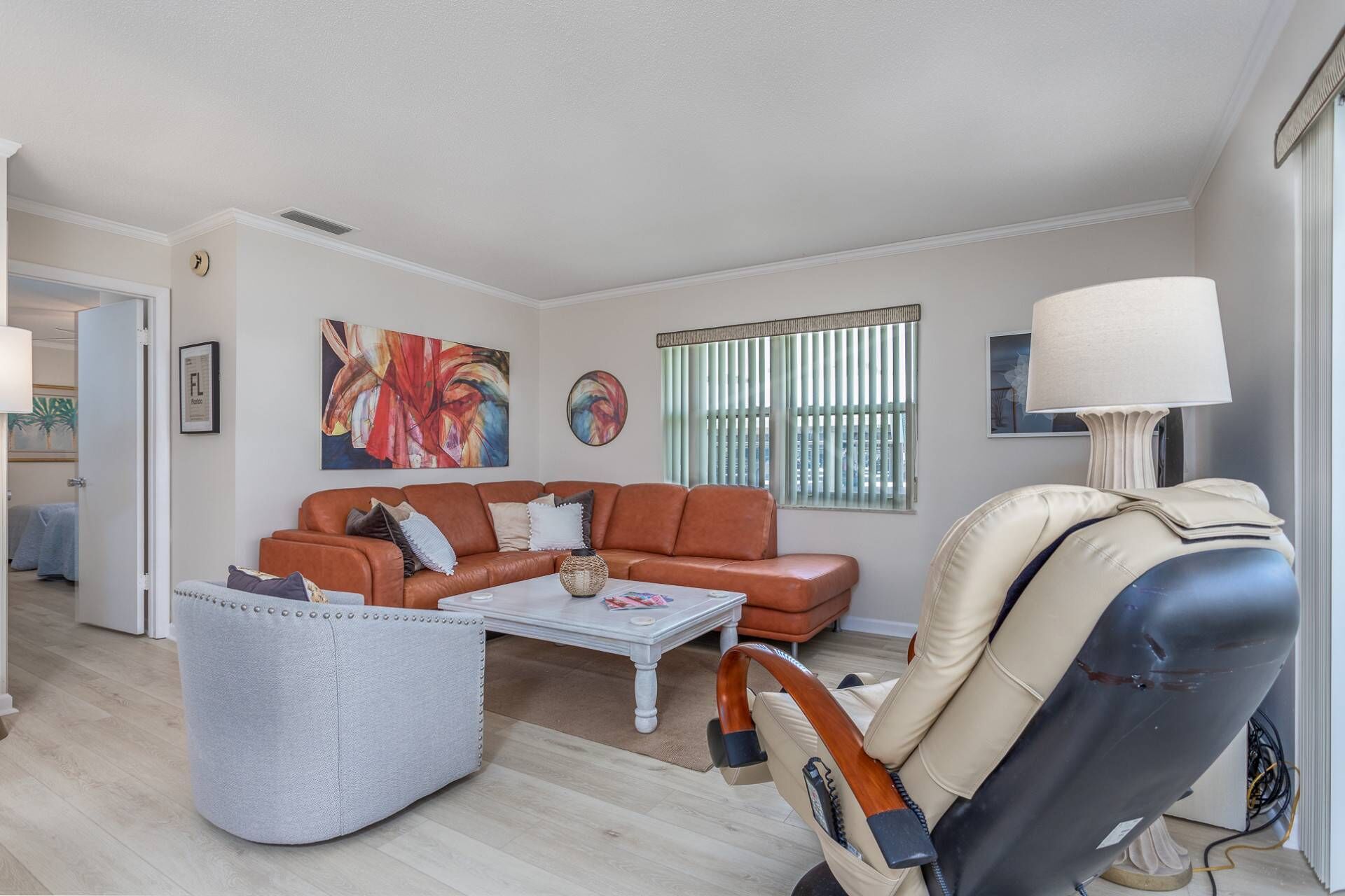 A living room with a brown leather sofa, a white coffee table, a grey pouf, a massage chair, and a large window with blinds.