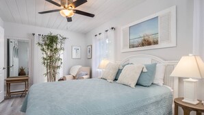 A bedroom with a bed, a ceiling fan, a window with curtains, a bedside table, and a lamp. - Seagrass Cottage (Sarasota)