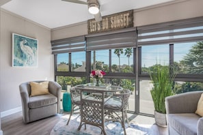 Relax in this inviting space with a view of palm trees and a residential area, featuring a comfortable sofa, dining table, and potted plant.