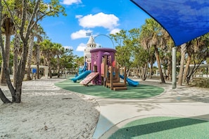 Kids can play and climb on the colorful play structure surrounded by a sandy beach and palm trees.