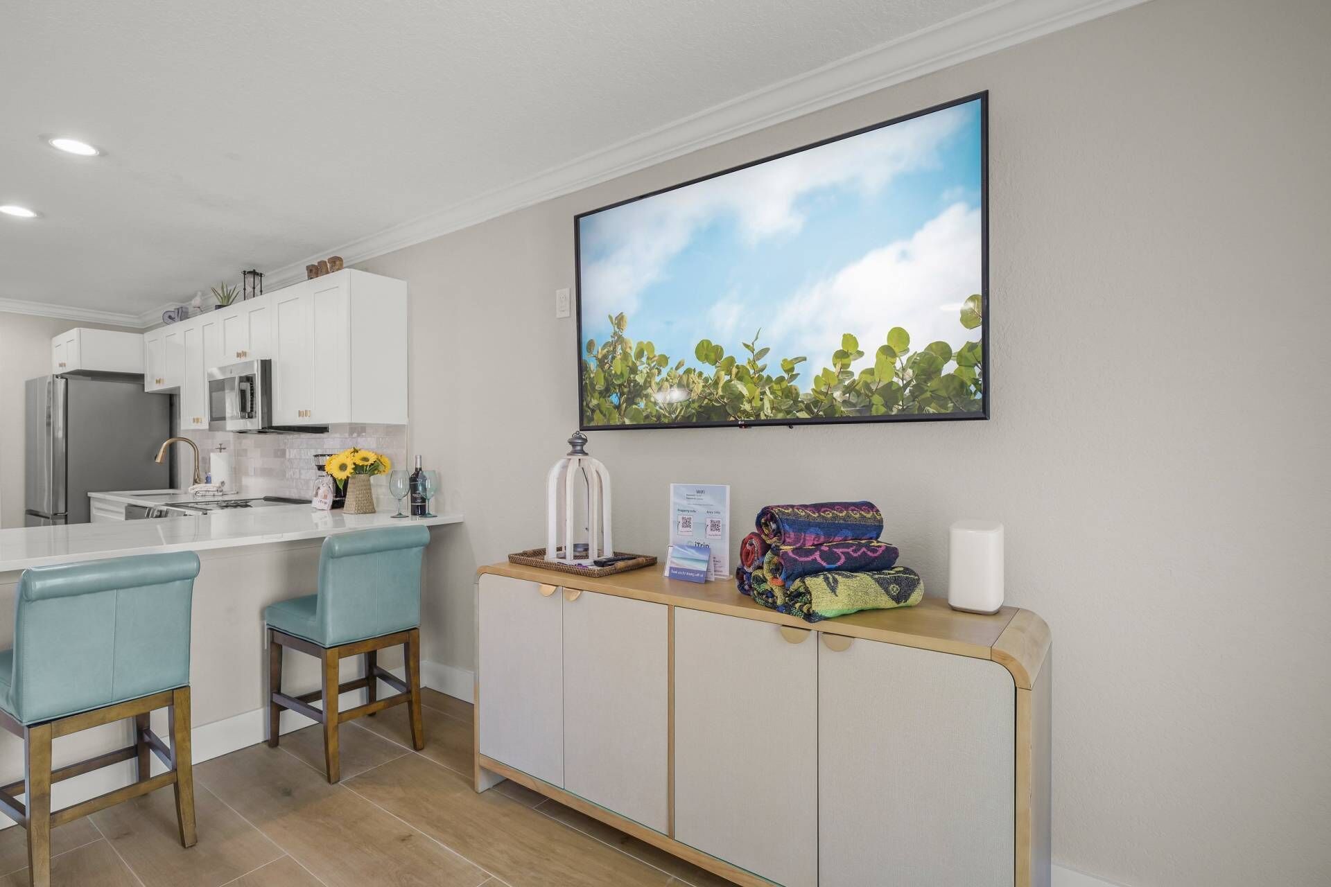 A modern kitchen with white cabinets, a stainless steel refrigerator, a microwave, and a sink. There are two blue bar stools and a wooden sideboard with a framed picture above it.