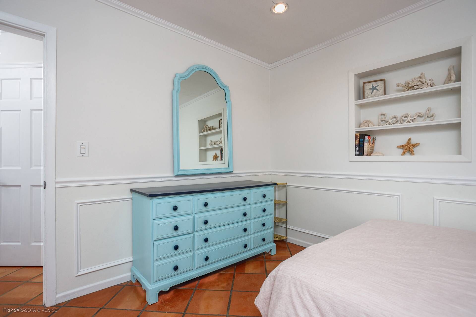 A bedroom with a light blue dresser, a large mirror, and a shelf with decorative items.