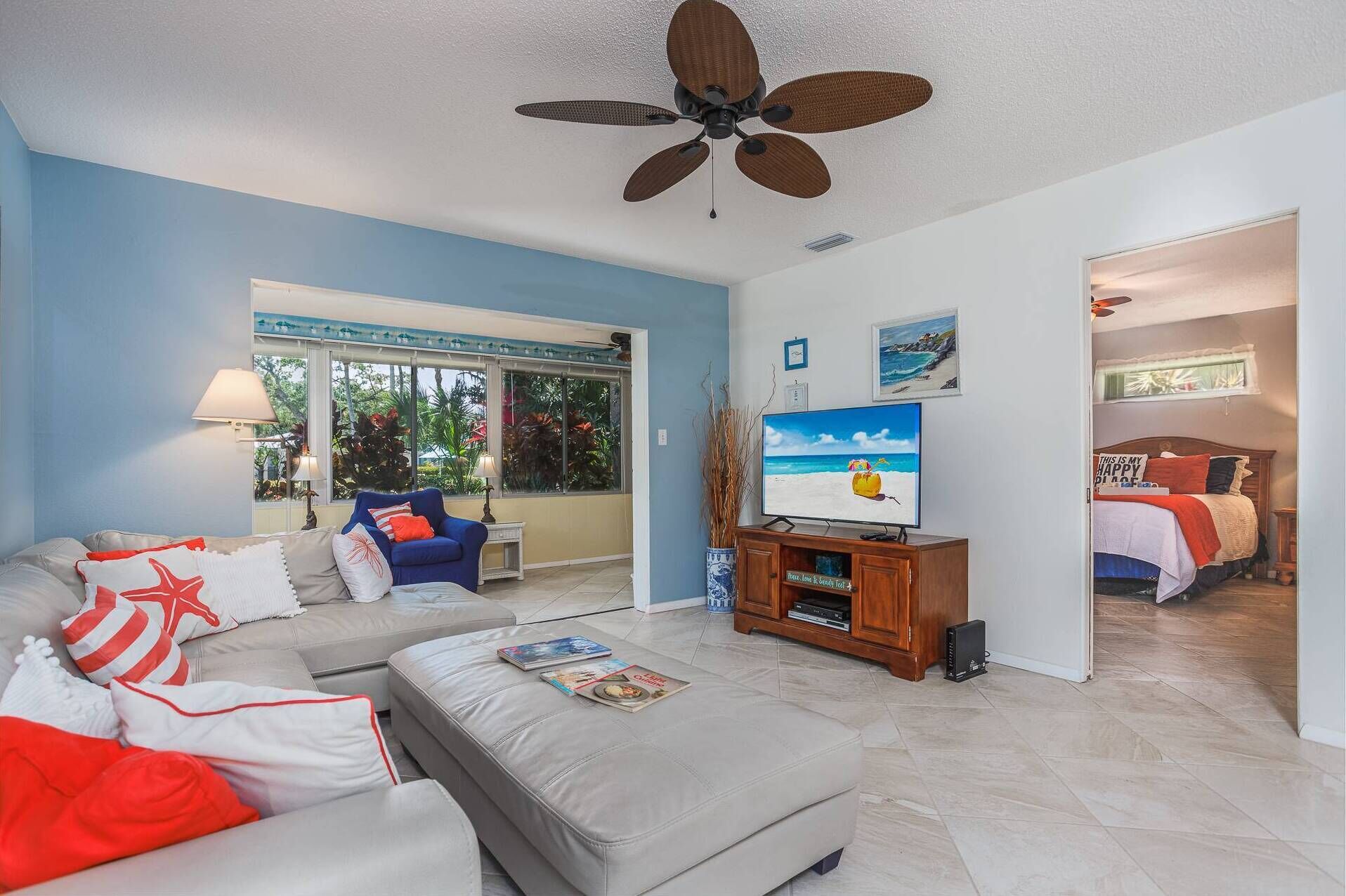 A living room with a large TV, a ceiling fan, a sofa, and a bed in the adjacent room.