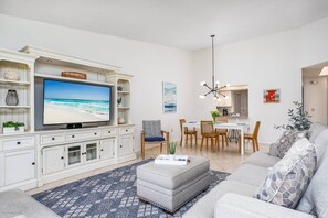 Een woonkamer met een flatscreen televisie, witte kasten, een grijze bank, een eethoek met een witte tafel en stoelen, en een keuken op de achtergrond.