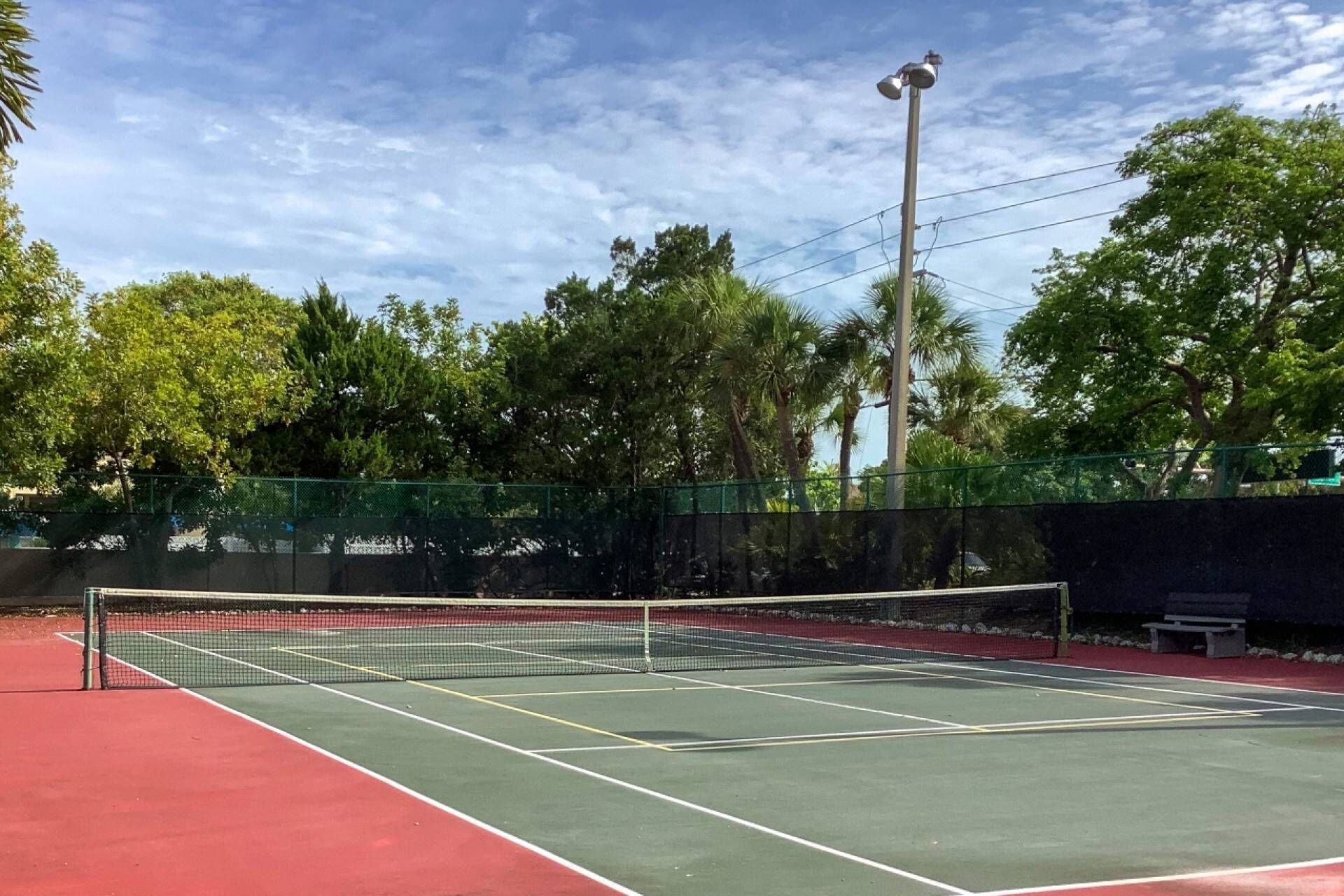 Ein Tennisplatz im Freien mit Netz, umgeben von einem grünen Zaun und Bäumen.