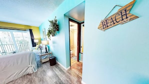 Une chambre d’hôtel avec un balcon, un lit, une table de chevet, une lampe et une salle de bain visible à travers une porte ouverte.
