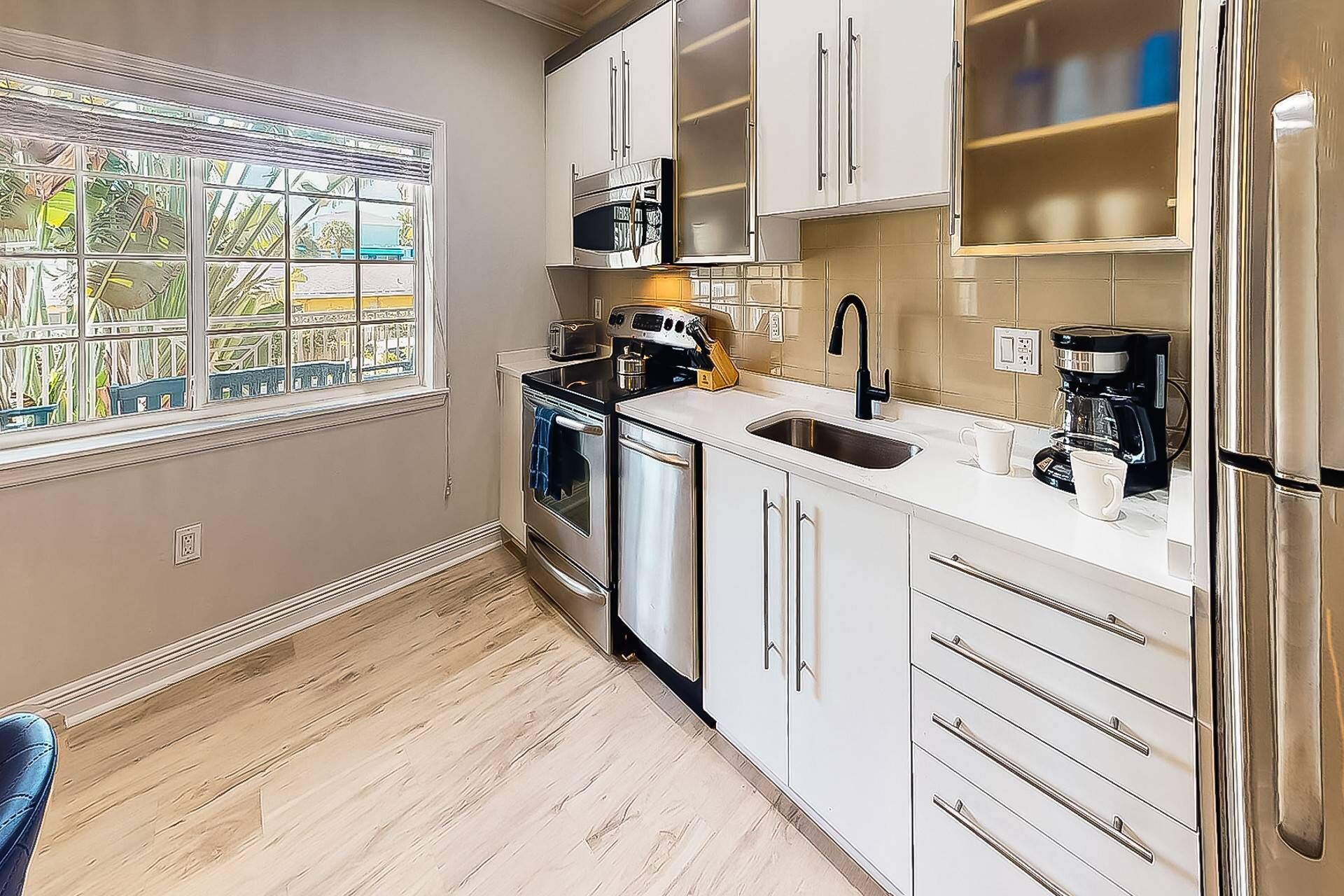 A modern kitchen with white cabinets, a stainless steel refrigerator, a coffee maker, and a window with a view of palm trees.