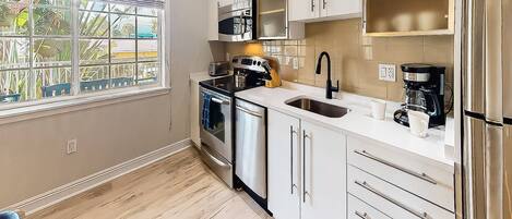 A modern kitchen with white cabinets, a stainless steel refrigerator, a coffee maker, and a window with a view of palm trees.