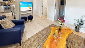 A modern living room with a blue sofa, a wooden coffee table with a vase and wine glasses, and a television displaying a beach scene. - Beach Club Modern Condo C Bldg (Siesta Key)