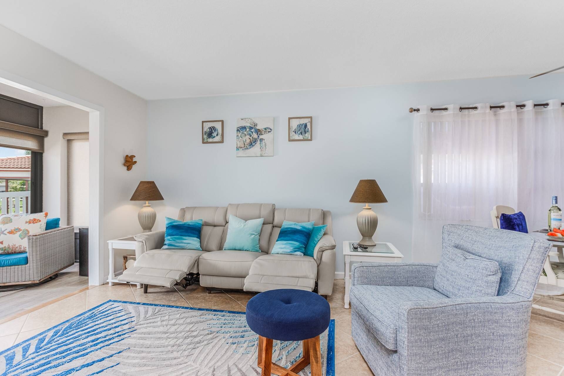 Un salon avec un canapé beige, un pouf bleu, un fauteuil gris et un tapis bleu. On y trouve deux lampes sur des tables d’appoint et trois œuvres d’art encadrées accrochées au mur.