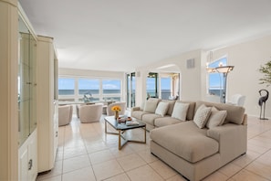 A spacious living room with a beige sofa, a coffee table, and a view of the sea through large windows.