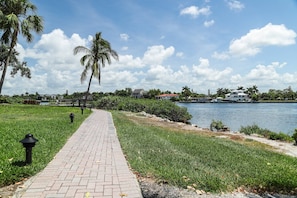 A paved walkway along a waterfront with palm trees and houses in the background. - Midnight Cove II Bldg 5, 1st Fl Close to Pool (Siesta Key)