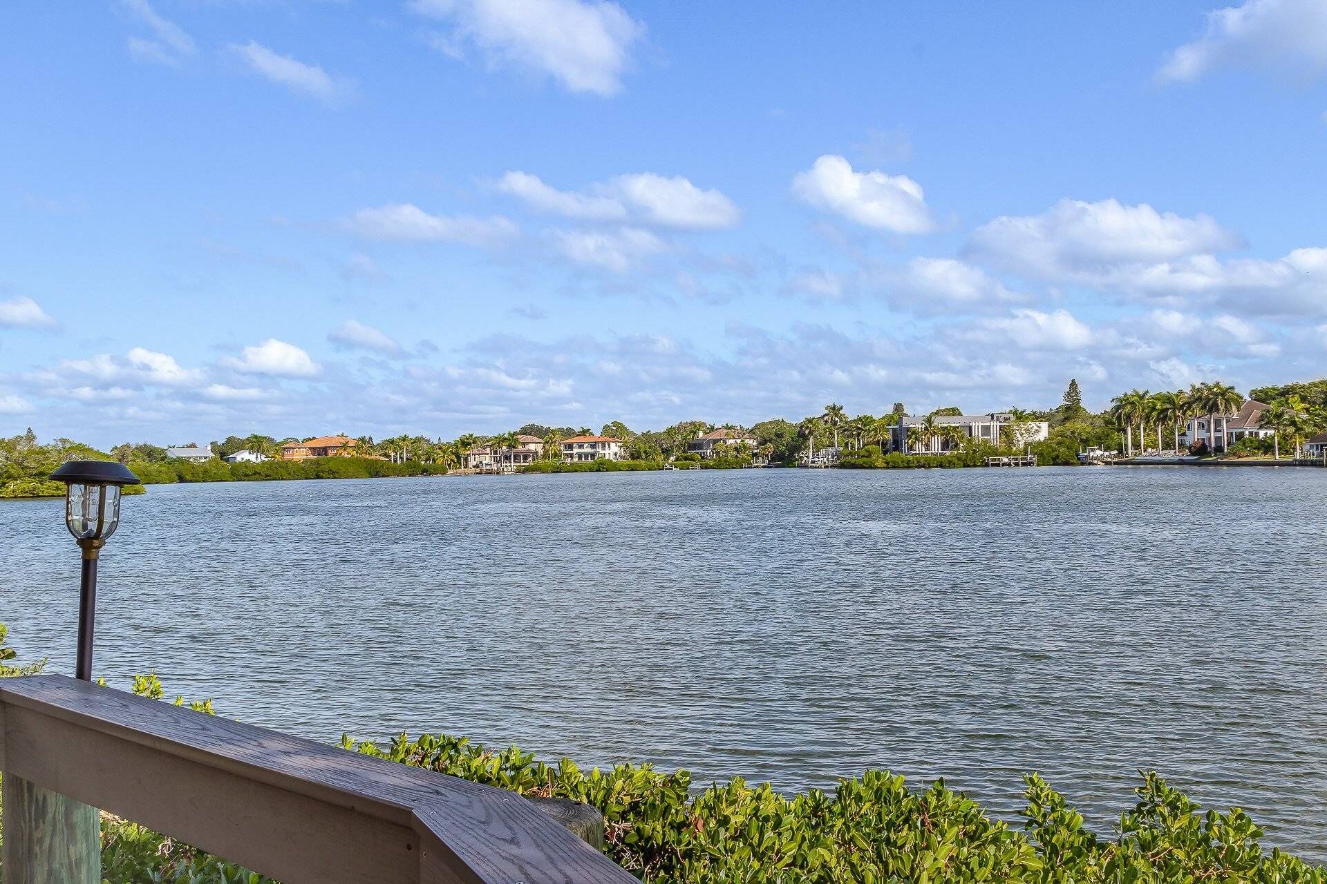 A waterfront view with a calm lake, a wooden deck, and a row of houses in the background.