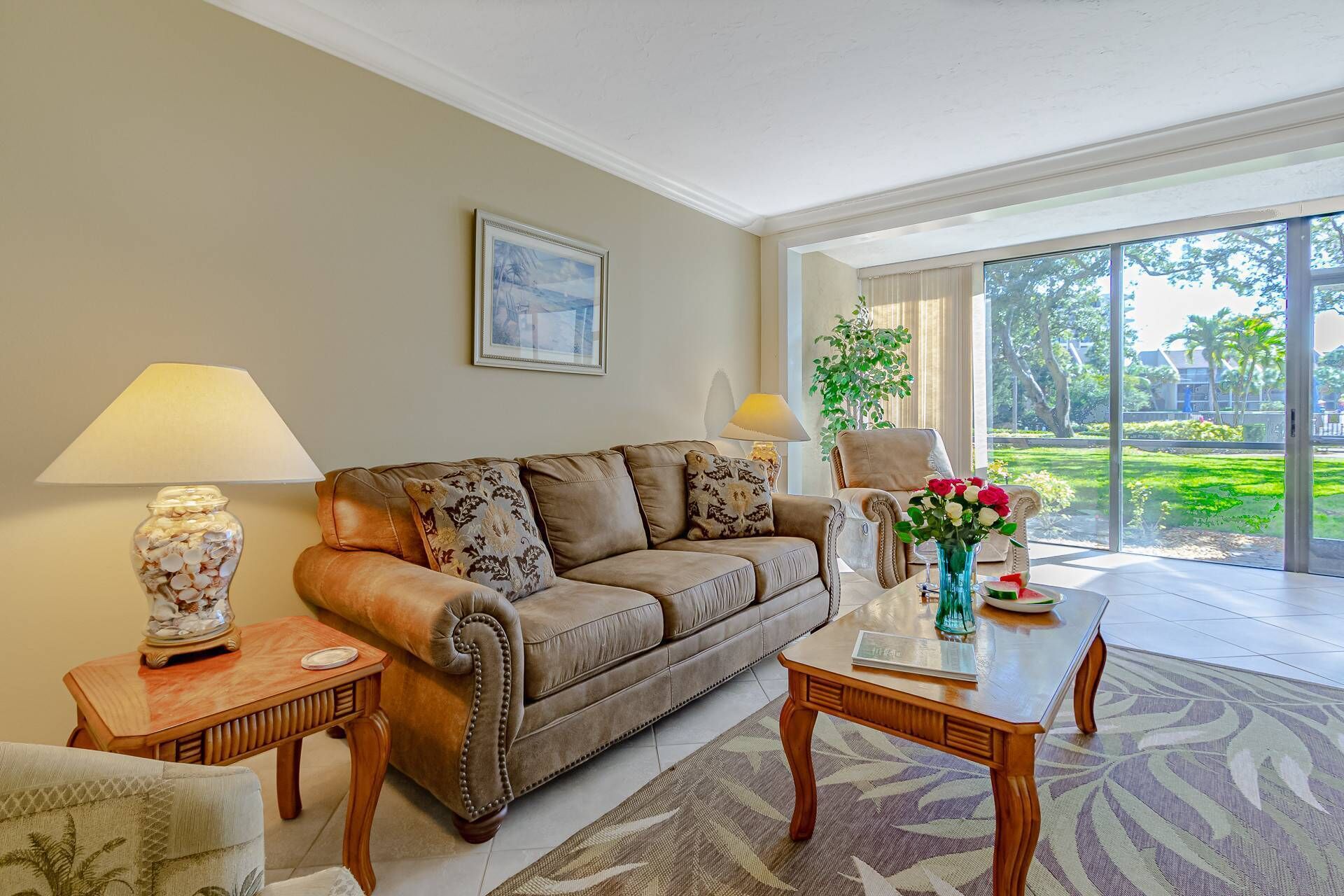 A living room with a brown leather sofa, two wooden side tables, a coffee table, and a large window with a view of greenery.