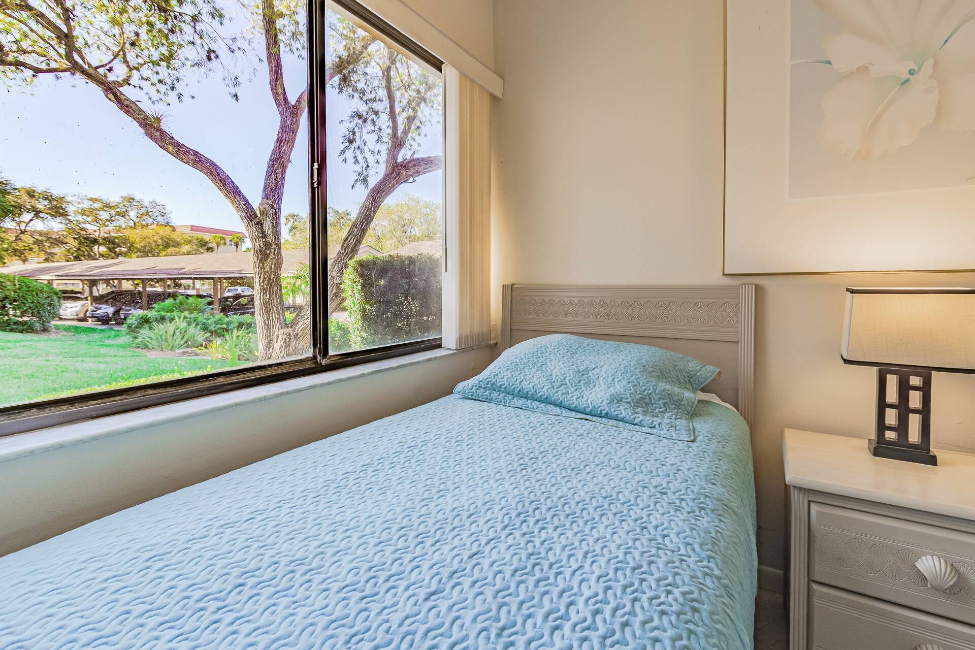 A bedroom with a bed, a nightstand, a lamp, and a window with a view of trees and a parking area.