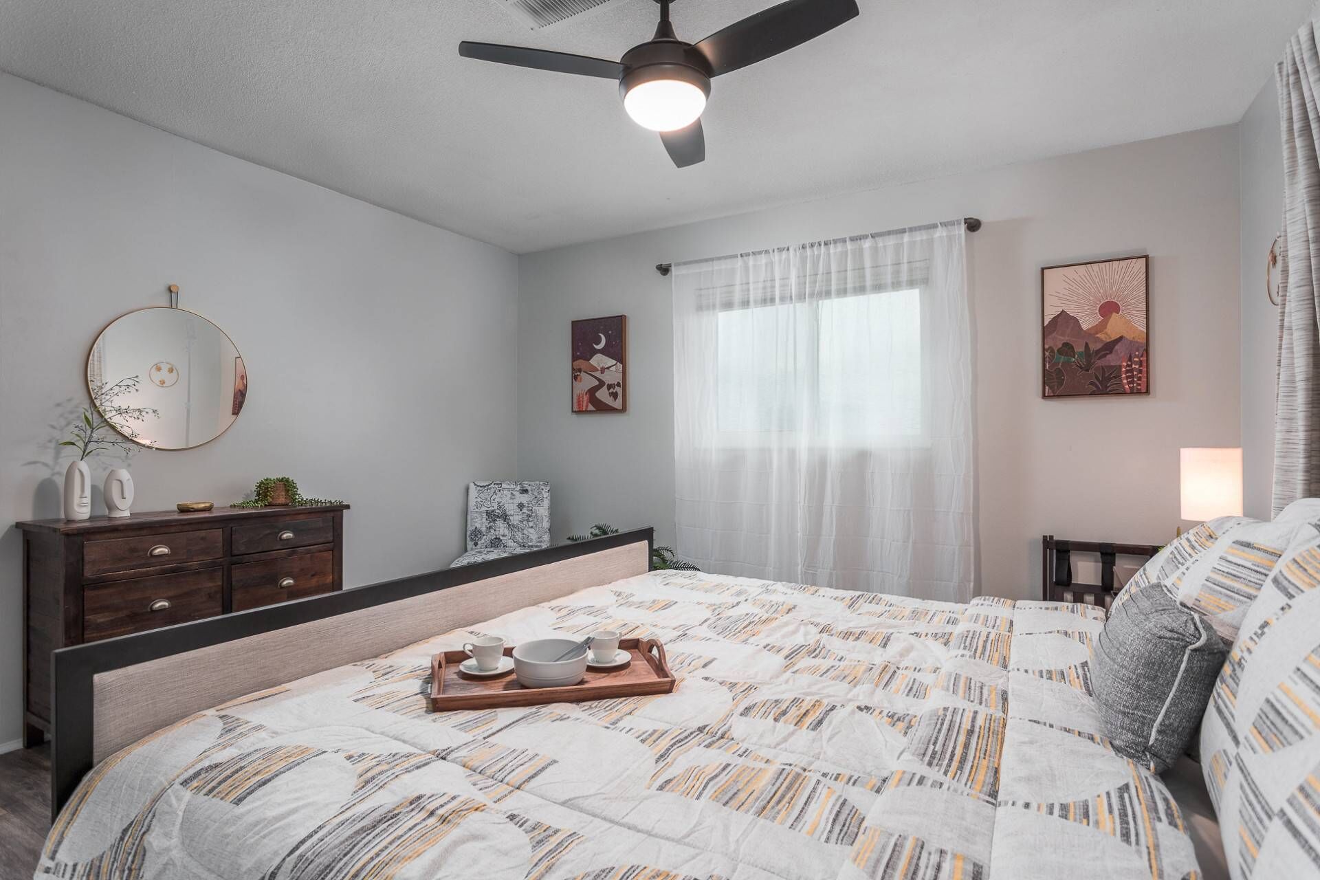 A bedroom with a bed, a dresser, a ceiling fan, a window with sheer curtains, and wall art.