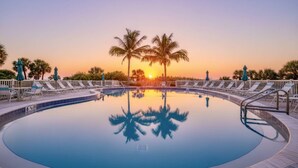 Une piscine circulaire entourée de chaises longues et de palmiers, dont l’eau reflète les couleurs du coucher de soleil.