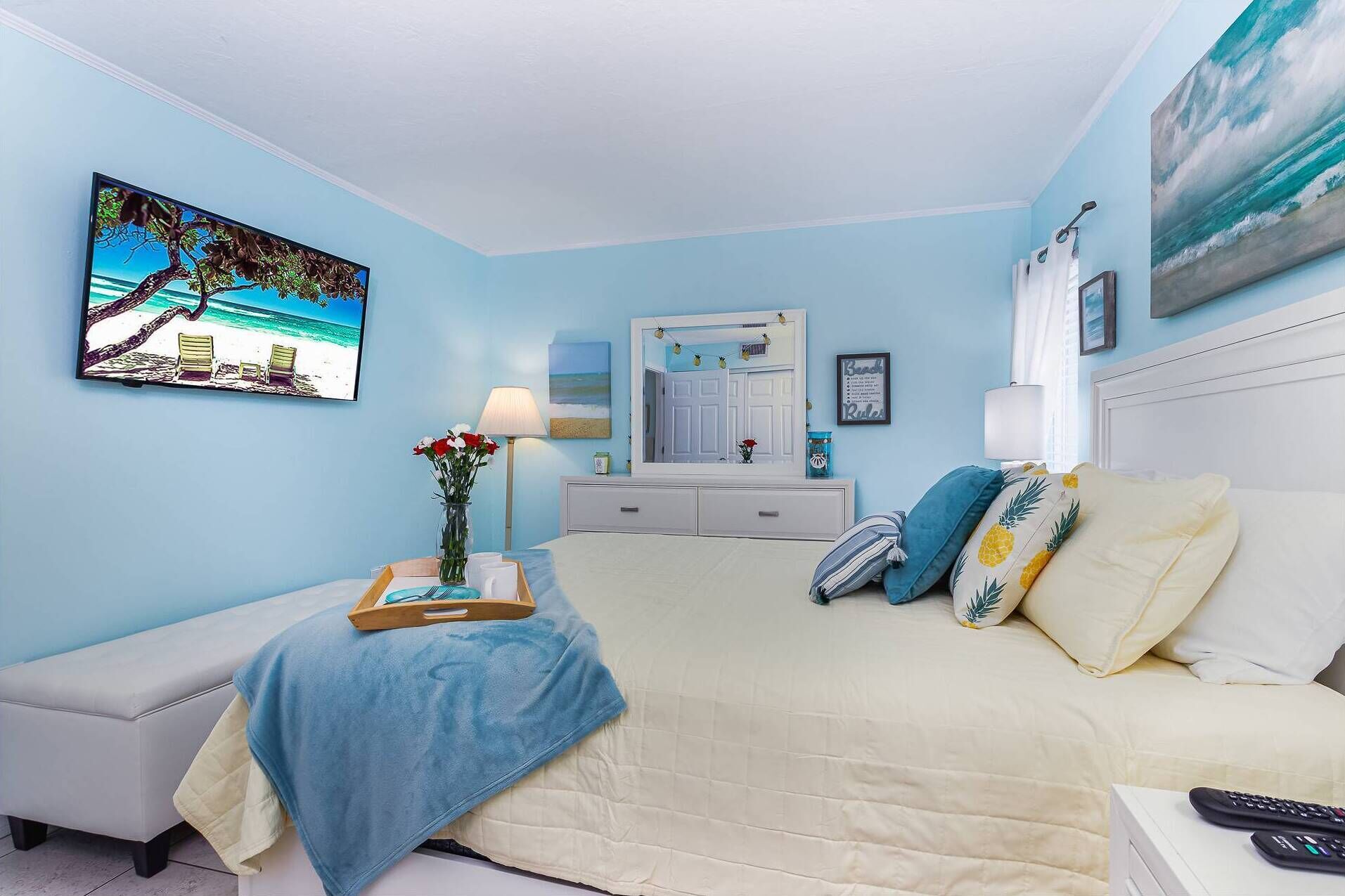 A bedroom with a bed, a bedside table with a tray, a lamp, a dresser, a mirror, and wall art.