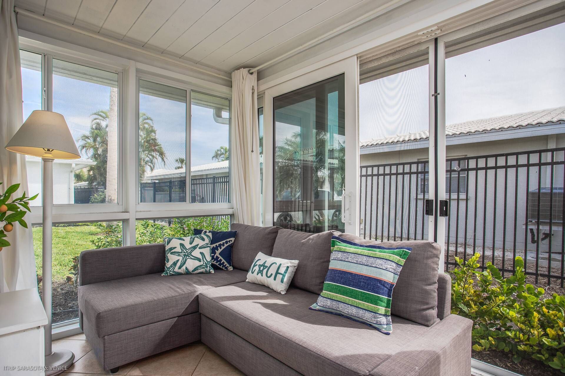 A corner sofa with patterned cushions, large windows, and a view of a fenced area outside.