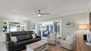 A spacious living room with a black leather sofa, a beige armchair, and a wooden coffee table. There is a ceiling fan, a painting of a turtle, and a lamp on a side table. - Crystal Sands & Seahorses Beach Villa (Siesta Key)