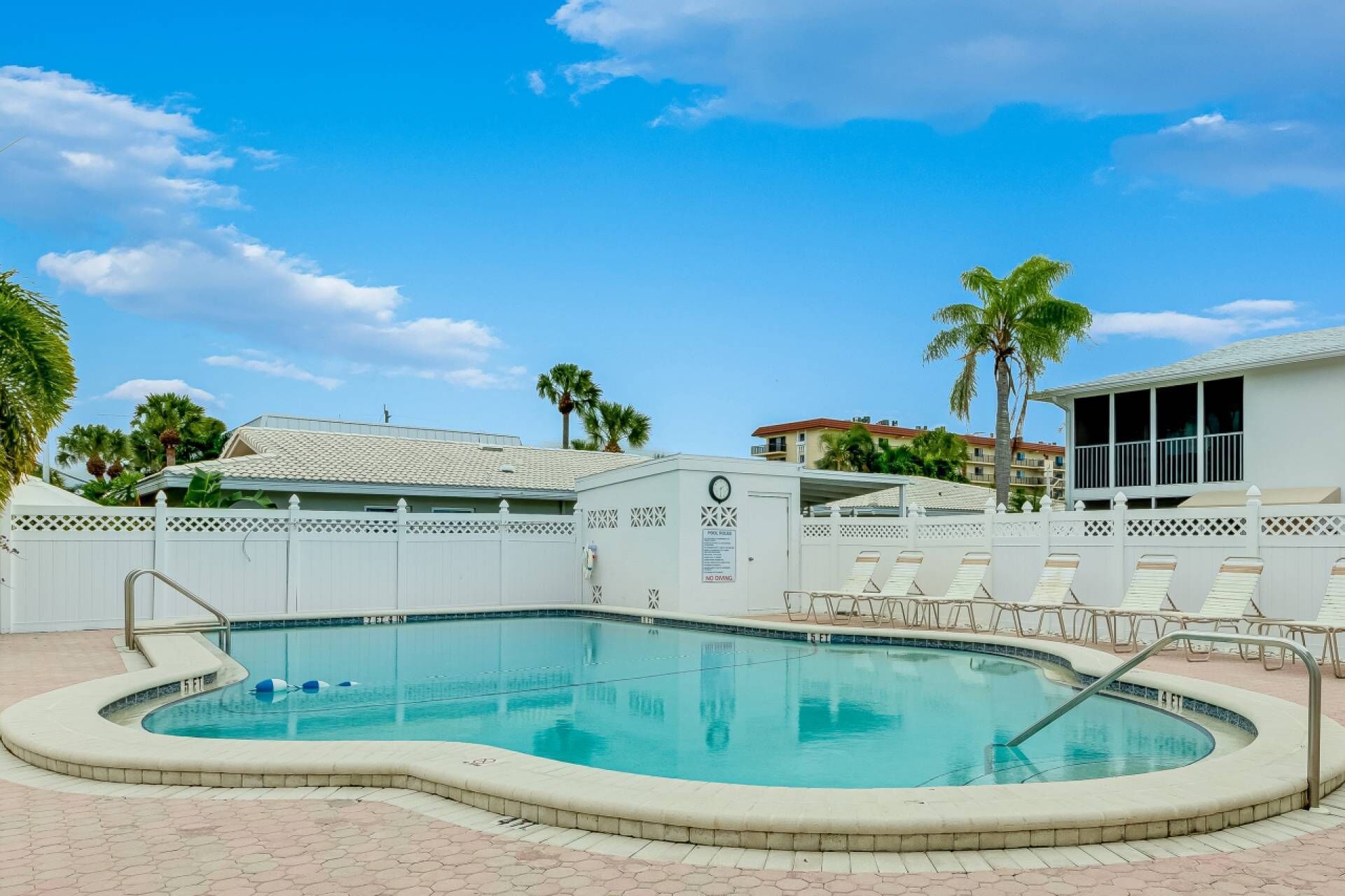 A swimming pool with a white fence, lounge chairs, and palm trees.