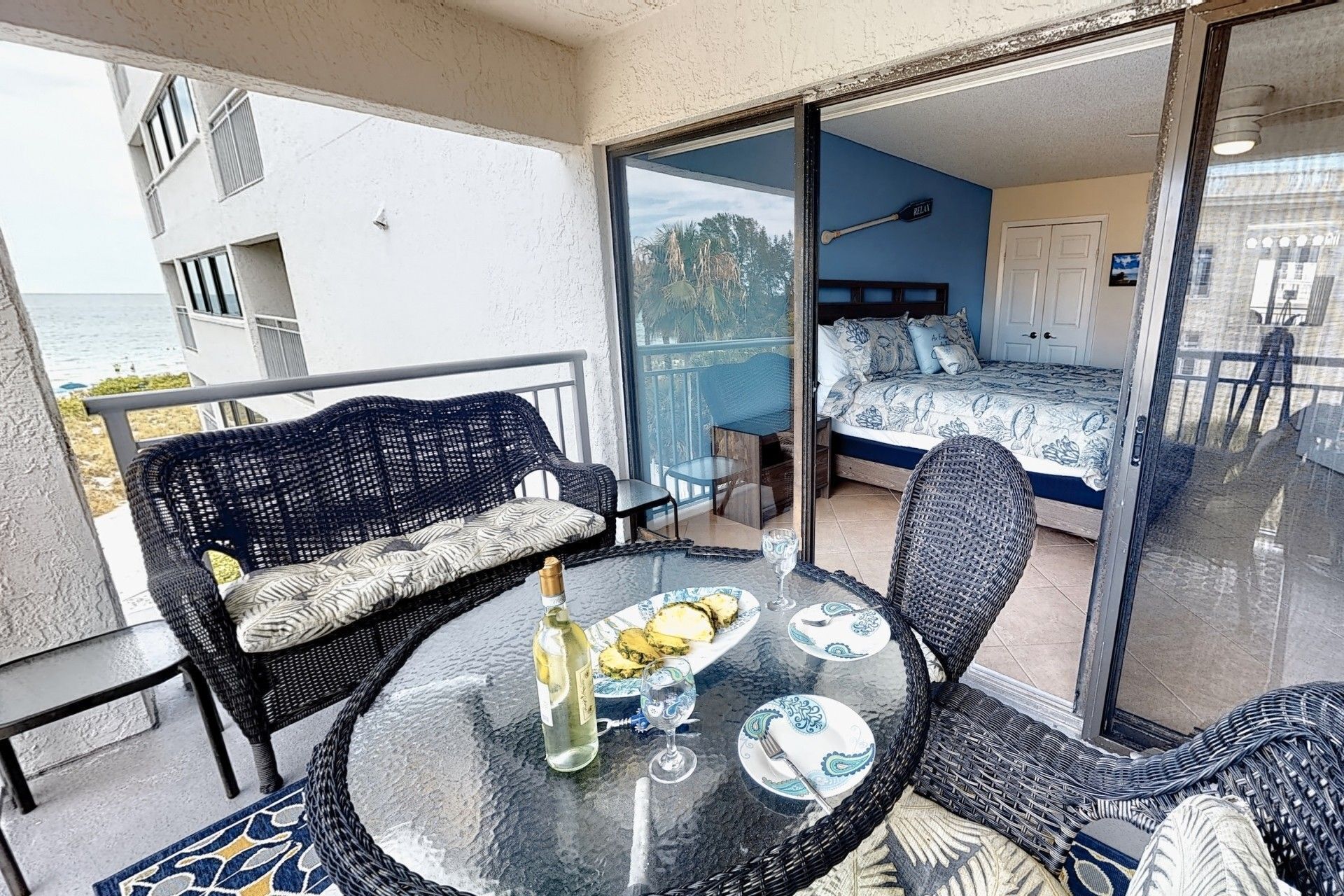 Een balkon met rieten meubilair, een glazen tafel en een bed met een blauw-witte spreidekker.
