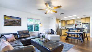 A modern living room with a sectional sofa, a coffee table, and a dining area with a bench and table. There is a kitchen in the background with wooden cabinets and stainless steel appliances. - Mova Poolside Escape (Sarasota)