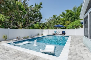 A rectangular swimming pool with a built-in waterfall feature, surrounded by a white fence, and two lounge chairs under a blue umbrella. - Mova Poolside Escape (Sarasota)