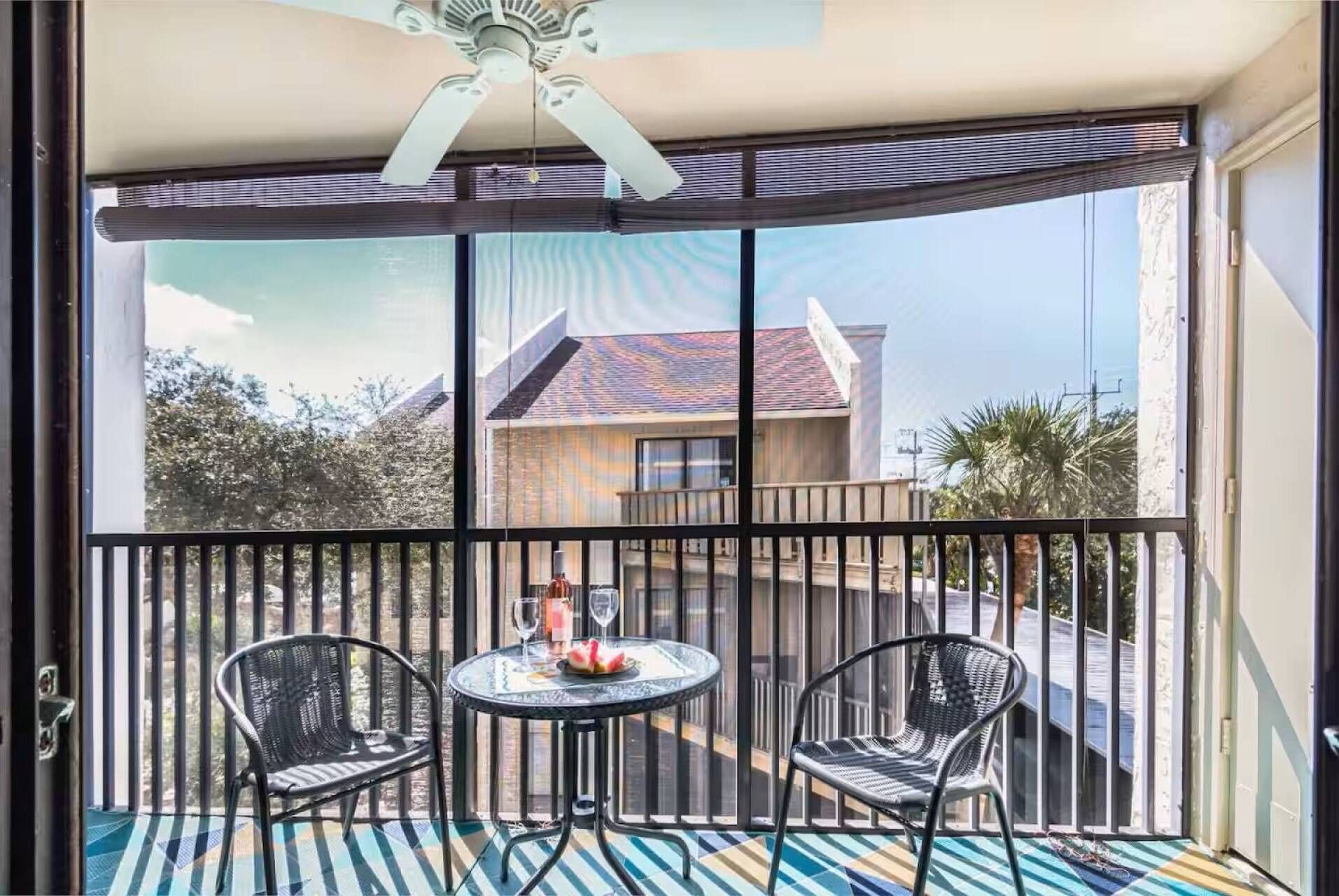 A balcony with a glass door, two wicker chairs, a small round table with a glass top, and a ceiling fan.