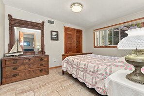 A bedroom with a wooden dresser, a bed with a floral bedspread, a wooden door, and a window with a view of the outdoors. - The Good Land Getaway (Moreno Valley)