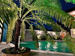 A pool area with a palm tree and a stone wall in the background. - Marmeu Tropical (Guarujá)