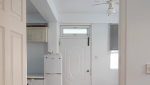 Ein schmales Entree mit einer weißen Tür, einer kleinen Küchenzeile mit Kühlschrank und einem Deckenventilator.