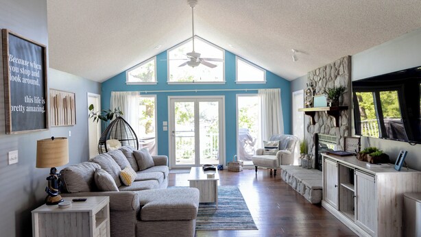 A spacious living room with a high ceiling, a ceiling fan, a large window, a fireplace, and a television.