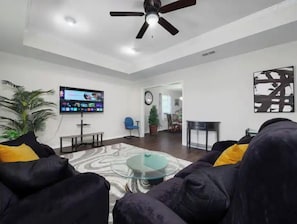 A living room with a flat-screen TV, ceiling fan, and a glass coffee table.