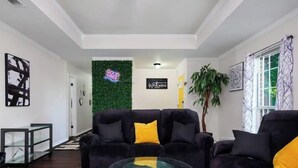 A modern living room with a glass coffee table, dark sofas with yellow cushions, a green wall with a 'Good Vibes' sign, and a 'Welcome' sign.