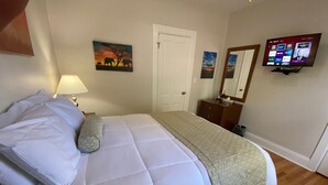 Een netjes opgemaakt bed met witte lakens en een bed met een geborduurd plaid, een bedlamp, een houten dressoir met spiegel, en een televisie waarop een streamingdienst draait.