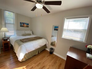 Une chambre à coucher comprenant un lit, un ventilateur de plafond, une table de chevet, une lampe, un miroir et une fenêtre avec des stores.
