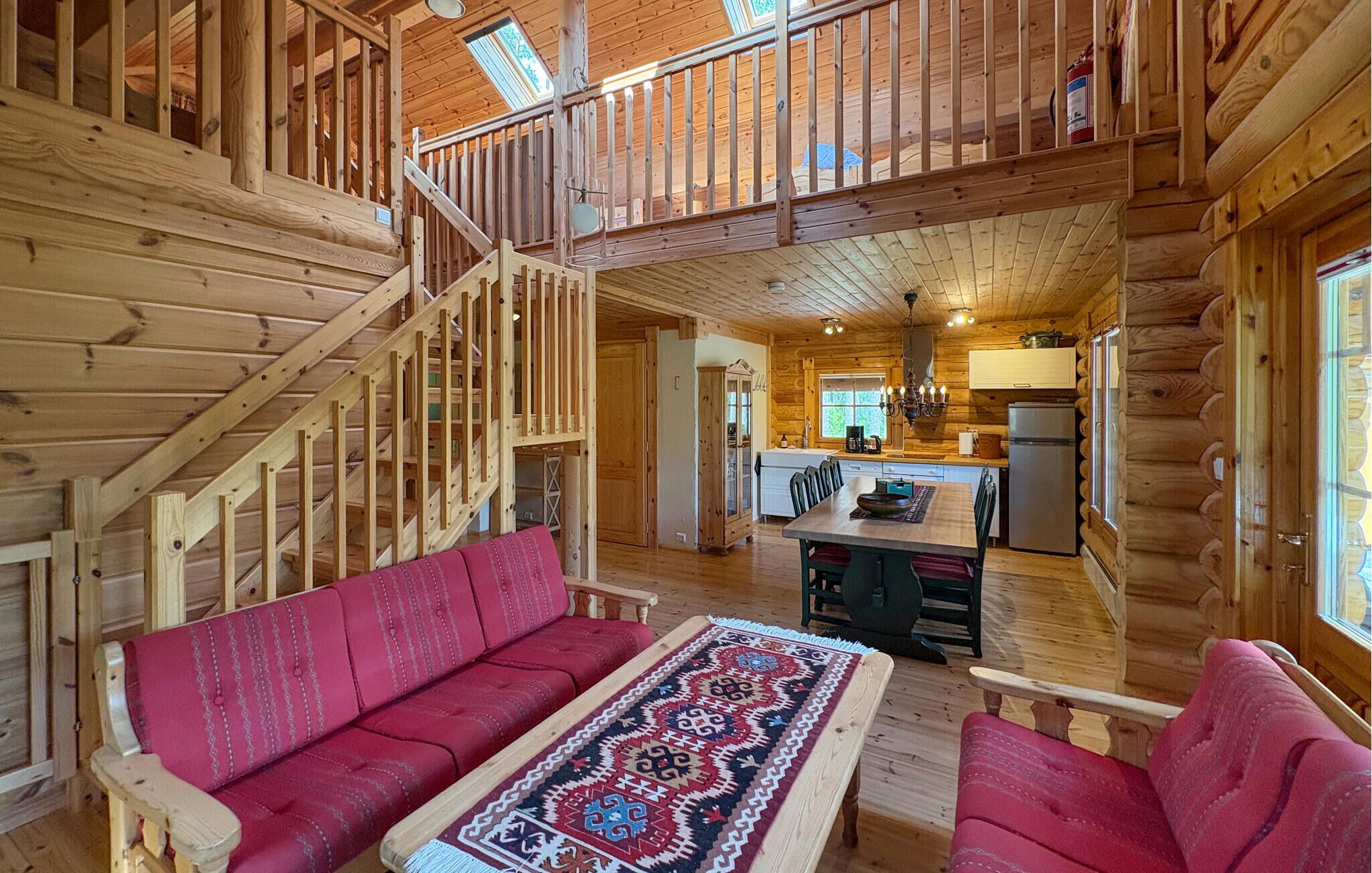 A log cabin interior with a wooden staircase, a dining area with a patterned rug, and a kitchen with a refrigerator.
