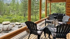 Eine Holzdiele mit Gartenmöbeln, darunter Stühle und ein Tisch, mit Blick auf eine bewaldete Gegend und dahinter Berge.