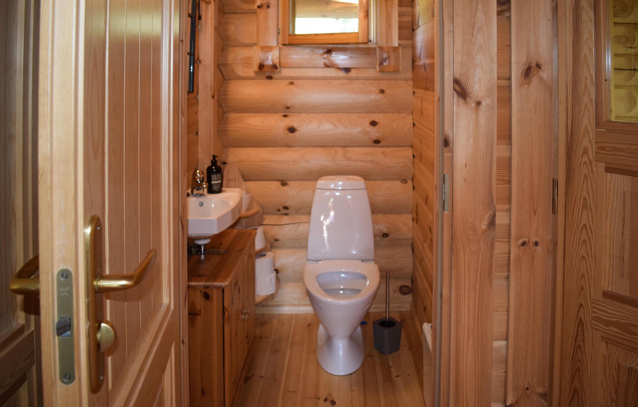 Baño de una cabaña de madera pequeña, con inodoro, lavamanos y una ventana pequeña sobre el lavamanos.
