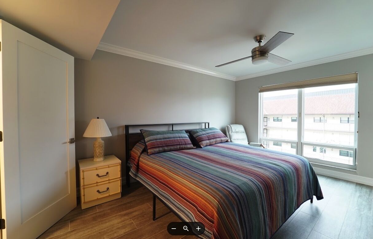 A bedroom with a bed, a nightstand, a lamp, a ceiling fan, and a window with a view of buildings.