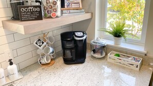 Eine Küche mit Kaffeemaschine, Tassen, einem Behälter für Kapseln, einem Kochbuch und einem Fenster mit Pflanze.