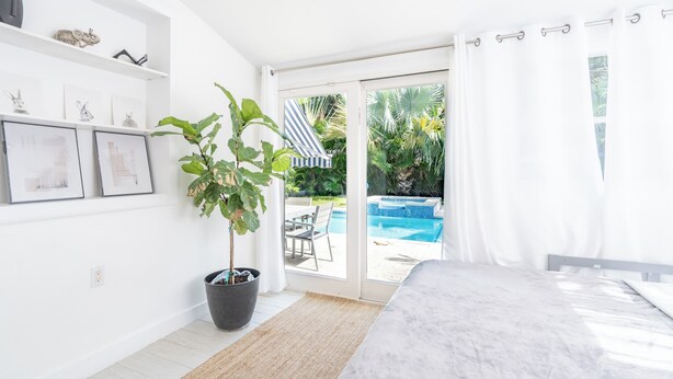 Un dormitorio con cama, planta en maceta, ventana con cortinas y vista a una piscina y zona de estar exterior.