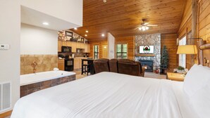 A spacious bedroom with a large bed, a wooden ceiling, a stone fireplace, and a kitchenette in the background.