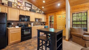 A kitchen with wooden cabinets, a black refrigerator, a microwave, and a stove. There is a wooden table with a stool, and a window with blinds.