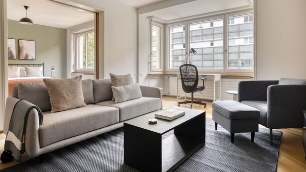 A modern living room with a grey sofa, a black coffee table, and a grey armchair. There is a large window with white frames and a view of buildings outside. - Sunny St. Alban  3 Room w/ Storage, nr Train Station, by Blueground (Basel)