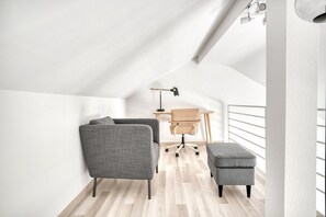 Quarto tipo loft moderno com teto alto, escrivaninha de madeira, cadeira de escritório cinza, sofá cinza e um banquinho cinza.