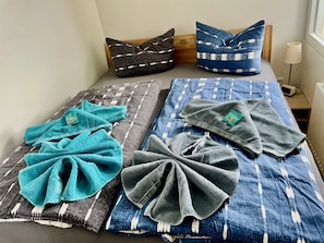 Una cama con una colcha azul y blanca estampada, dos almohadas con diseños negros y blancos, y dos toallas dobladas en tonos verde azulado y gris.